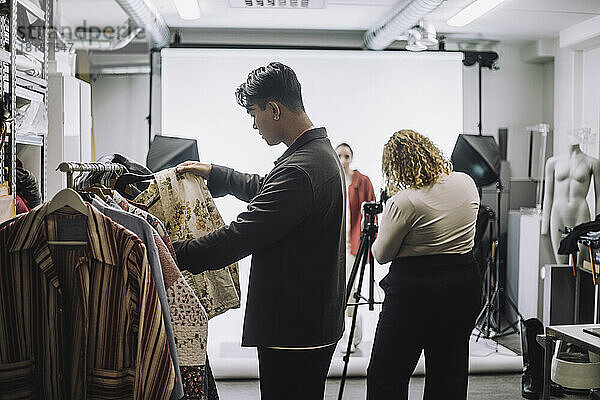 Seitenansicht eines männlichen Designers  der während eines Fotoshootings im Studio Kleidung von der Stange auswählt