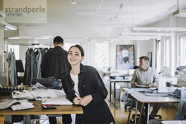 Porträt einer lächelnden jungen Modedesignerin  die sich in einer Werkstatt an eine Werkbank lehnt