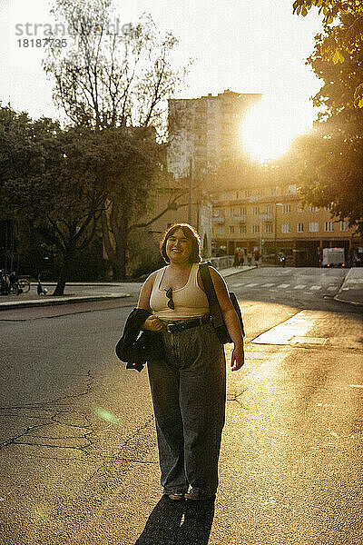 Porträt einer lächelnden jungen Frau  die bei Sonnenuntergang auf der Straße steht