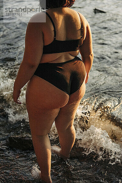 Tiefschnitt einer jungen Frau in einem schwarzen Bikini  die am Strand steht und im Wasser plantscht