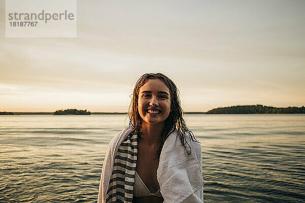 Porträt einer glücklichen Frau mit Handtuch am See bei Sonnenuntergang
