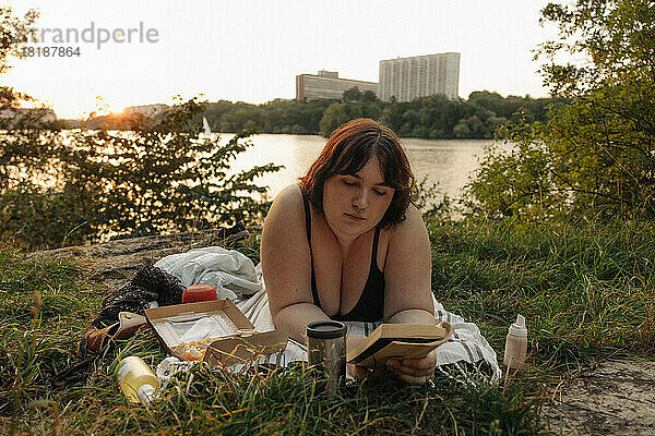 Junge Frau liest ein Buch  während sie bei Sonnenuntergang am See liegt