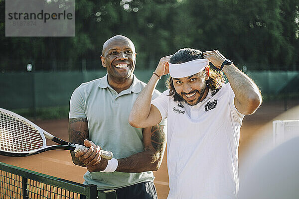 Glücklicher Mann mit Tennisschläger und männlichem Freund  der ein Stirnband auf dem Sportplatz einstellt