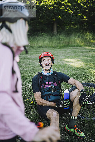 Glücklicher männlicher Radfahrer sitzt im Gras und hält eine Wasserflasche  während er mit einer Freundin spricht