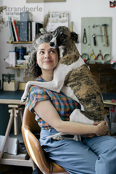 Lächelnde Frau  die ihren Hund umarmt  während sie zu Hause auf einem Stuhl sitzt