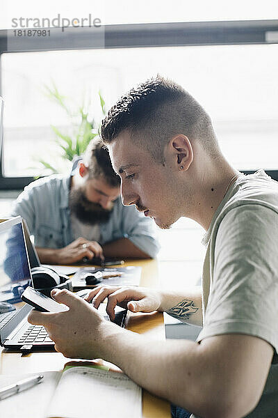 Männlicher Computerprogrammierer  der ein Smartphone neben einem Laptop auf einem Schreibtisch in einem Startup-Unternehmen benutzt