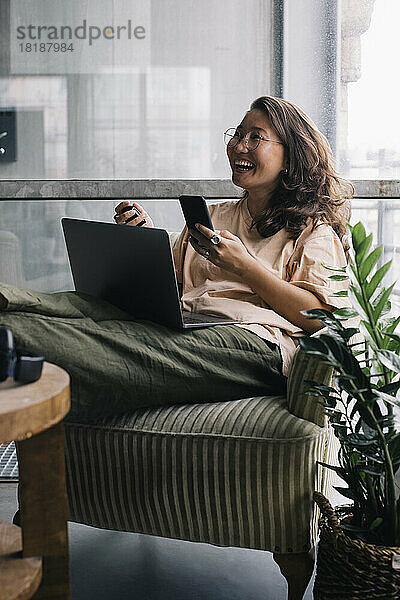 Weiblicher Computerprogrammierer mit Laptop und Smartphone lachend im Büro