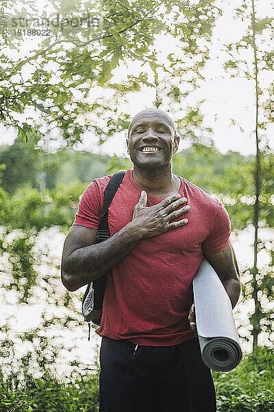 Lächelnder Mann mit Hand auf der Brust  der eine Übungsmatte im Wald hält