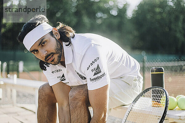 Selbstbewusster Mann mit Stirnband  der auf einer Bank auf dem Tennisplatz sitzt