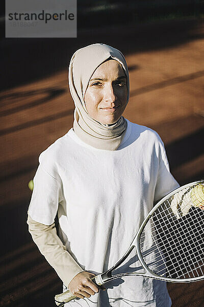 Selbstbewusste Frau mit Kopftuch hält Tennisschläger an einem sonnigen Tag