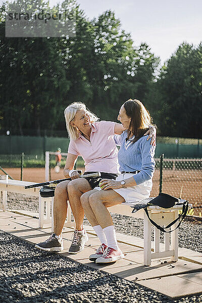 Glückliche ältere Freundinnen  die sich auf einer Bank am Tennisplatz unterhalten