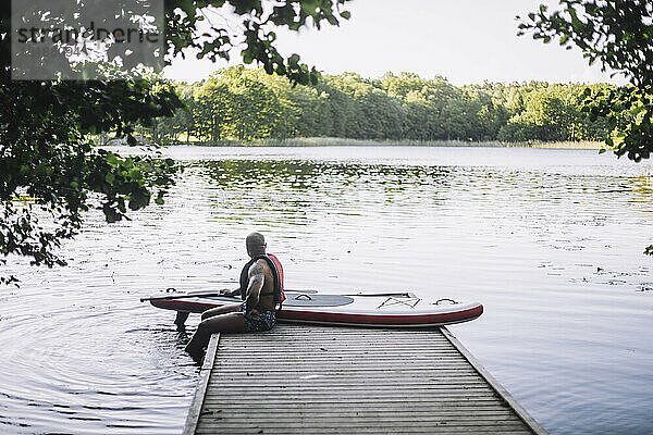 Mann mit Paddelboard schaut auf den See  während er auf dem Steg sitzt