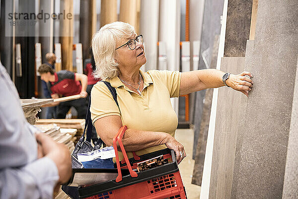 Ältere Frau trägt einen Einkaufskorb und wählt im Baumarkt laminierte Platten aus