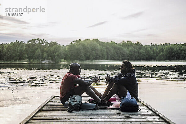 Mann teilt sein Smartphone mit einem Freund  der auf einem Steg am See sitzt