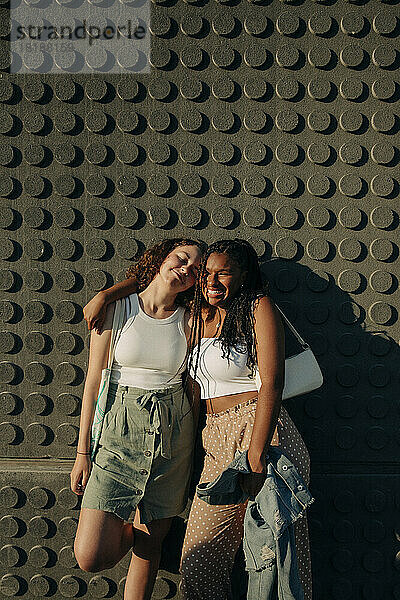 Porträt eines glücklichen Teenager-Mädchens  das mit einem Arm um einen Freund an der Wand steht  während eines sonnigen Tages