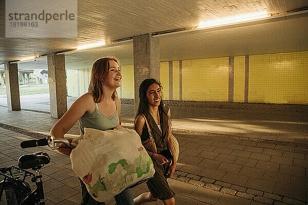 Lächelndes Teenager-Mädchen mit Fahrrad und Freundin unter einer Brücke