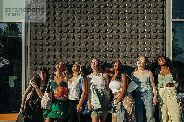 Lächelnde Teenager-Mädchen mit geschlossenen Augen genießen den sonnigen Tag  während sie an der Wand stehen