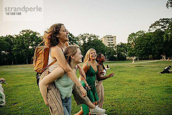 Glückliches Mädchen im Teenageralter  das seine Freundin beim Huckepackfahren im Park beobachtet