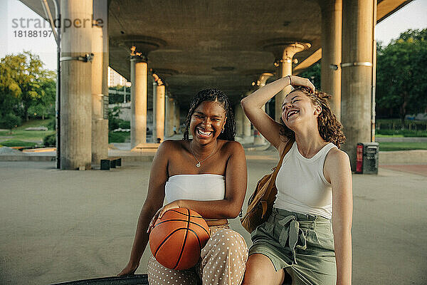 Fröhliches Teenager-Mädchen mit Basketball  das mit seiner Freundin unter einer Brücke sitzt