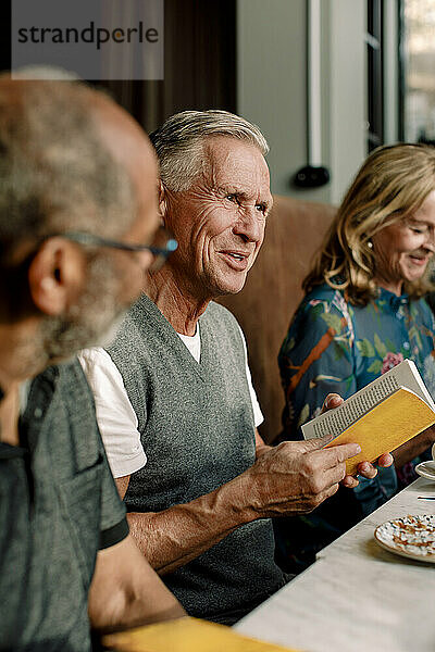 Älterer Mann hält ein Buch in der Hand und unterhält sich mit männlichen und weiblichen Freunden in einem Café