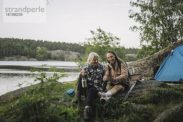 Ältere Freundinnen teilen ihr Smartphone  während sie beim Camping auf einem Felsen sitzen