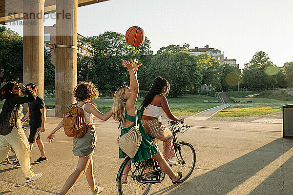 Rückansicht eines jugendlichen Mädchens  das einen Basketball wirft  während es mit einer Freundin auf dem Fahrrad im Park sitzt