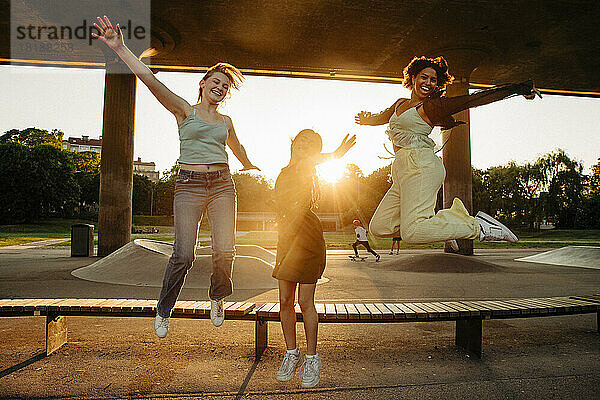 Porträt von glücklichen Teenager-Mädchen mit ausgestreckten Armen  die während des Sonnenuntergangs auf einer Bank springen
