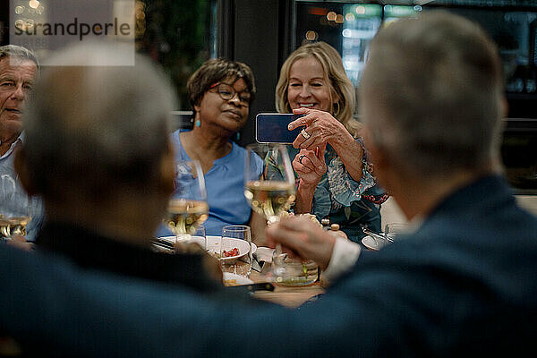 Lächelnde ältere Frauen fotografieren männliche Freunde mit Weingläsern in einem Restaurant