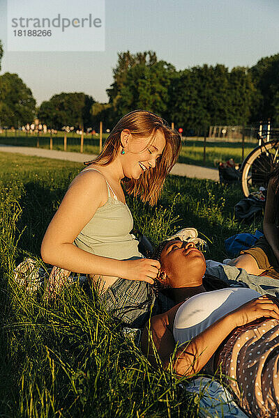 Lächelndes Teenager-Mädchen im Gespräch mit einer auf dem Schoß liegenden Freundin im Park