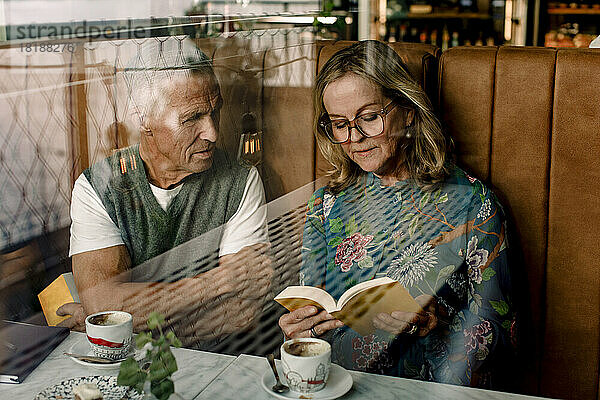 Ältere Frau liest einem männlichen Freund ein Buch vor  der mit verschränkten Armen in einem Café sitzt  gesehen durch Glas