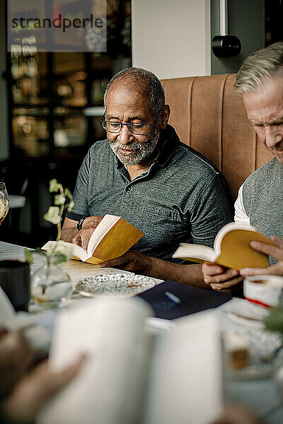 Älterer Mann mit Brille liest ein Buch  während er neben einem männlichen Freund in einem Café sitzt
