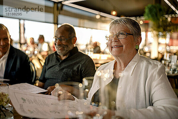 Lächelnde ältere Frau mit Brille sitzt mit Menükarte bei Männern im Restaurant