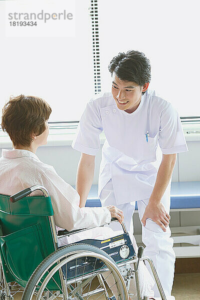 Eine japanische Seniorin im Rollstuhl und ein junger Krankenpfleger unterhalten sich