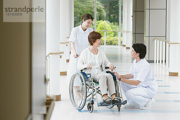 Eine japanische Seniorin im Rollstuhl und zwei junge Krankenschwestern unterhalten sich auf dem Flur