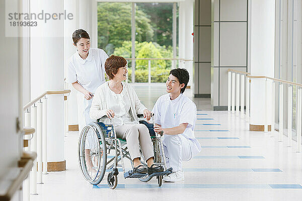 Eine japanische Seniorin im Rollstuhl und zwei junge Krankenschwestern unterhalten sich auf dem Flur