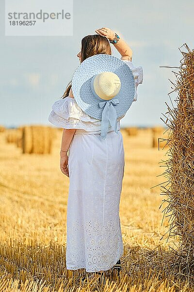 Rückansicht einer Frau in weißem Sommerkleid und Strohhut  die an einem sonnigen Tag in der Nähe eines Heuhaufens auf dem Feld steht