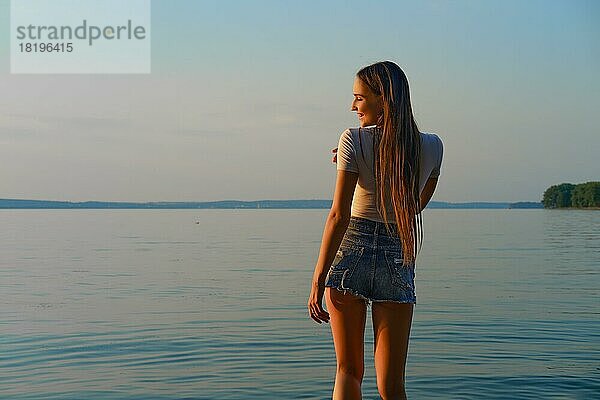 Hübsche Frau in Jeans-Shorts und weißem Unterhemd steht am Wasser in einem Erholungsgebiet bei Sonnenaufgang