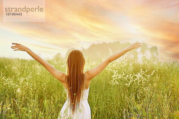 Junge Frau  die ihre Hände seitlich ausbreitet und der Sonne zugewandt auf einem Feld steht