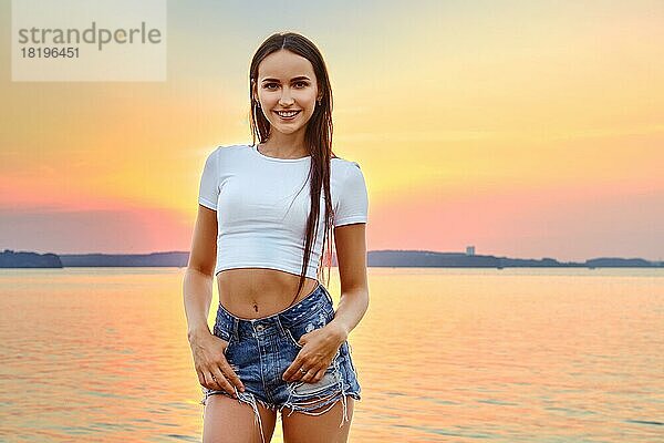 Fröhliche Frau in weißem T-Shirt und Jeansshorts posiert auf einem Pier mit Blick auf den herrlichen Sonnenuntergang