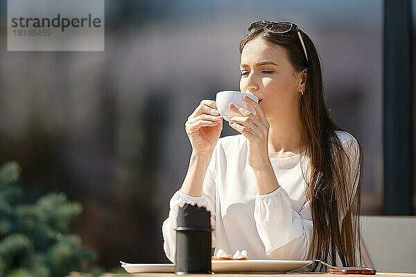 Unbekümmerte junge Frau sitzt auf der Terrasse eines Cafés und trinkt Cappuccino