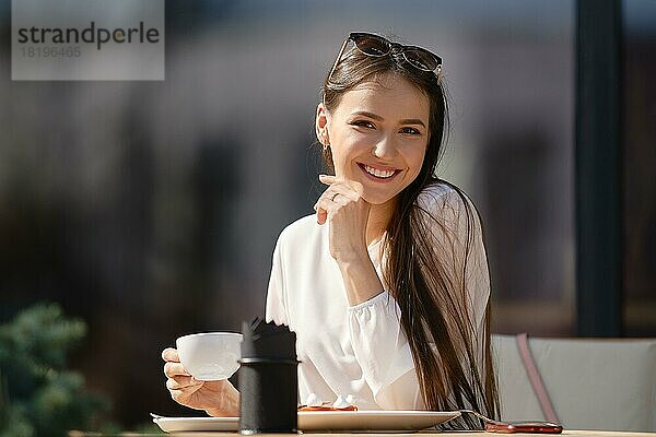 Hübsche junge Frau mit einer Tasse Cappuccino in der Hand schaut geradeaus und lächelt