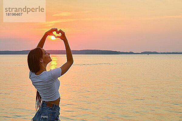 Junge Frau zeigt Liebeszeichen mit ihren Armen. Sie legte die Finger in Form eines Herzens in Richtung Sonnenuntergang