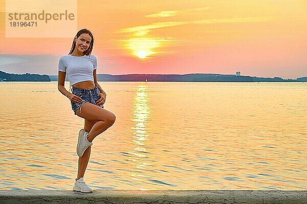 Fröhliche Frau in T-Shirt und Jeansshorts posiert auf einem Pier bei Sonnenuntergang