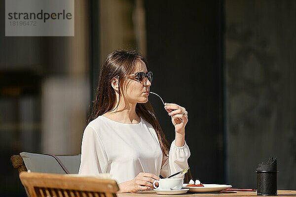 Schöne junge Frau mit Sonnenbrille sitzt auf der Terrasse des Cafés in sonnigen Tag und essen Kuchen