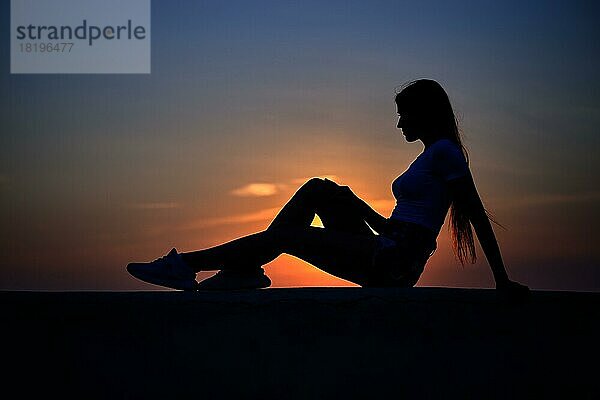 Silhouette einer im Profil sitzenden Frau auf einem Pier während des Sonnenuntergangs