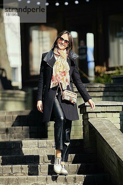 Fröhliche junge Frau in Mantel und Lederhose beim Hinabsteigen der Treppe in der Stadt