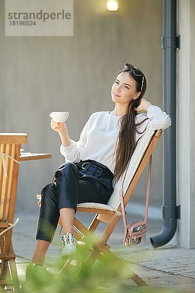 Glückliche junge Frau sitzt auf der Terrasse des Cafés in sonnigen Tag und trinken Kaffee. Frau genießt erste Frühlingssonne im Freien
