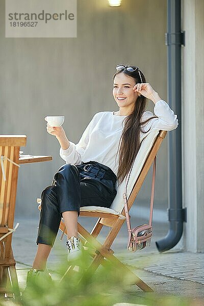 Glückliche junge Frau sitzt auf der Terrasse des Cafés in sonnigen Tag und trinken Kaffee. Frau genießt erste Frühlingssonne im Freien