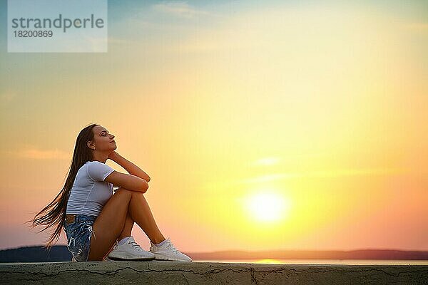 Ruhige und verträumte Frau sitzt im Profil auf einem Pier und genießt den Sonnenuntergang