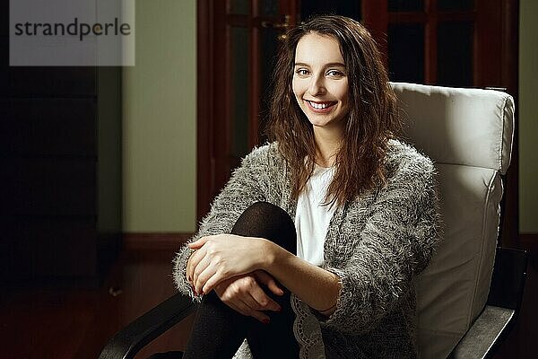 Glückliches lächelndes Mädchen sitzt in einem Sessel im Zimmer und schaut geradeaus